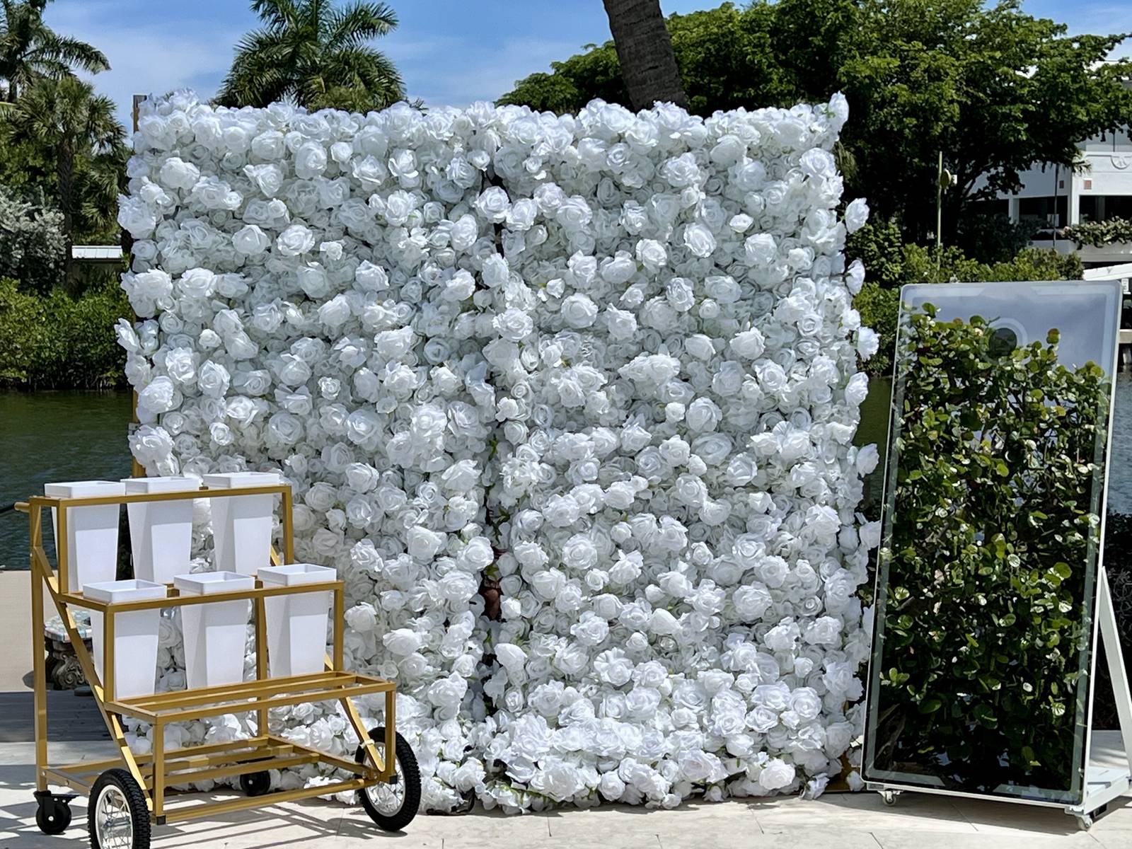 The white rose flower wall with the gold Bloom Bar cart and the mirror booth beside it, on a terrace by the water